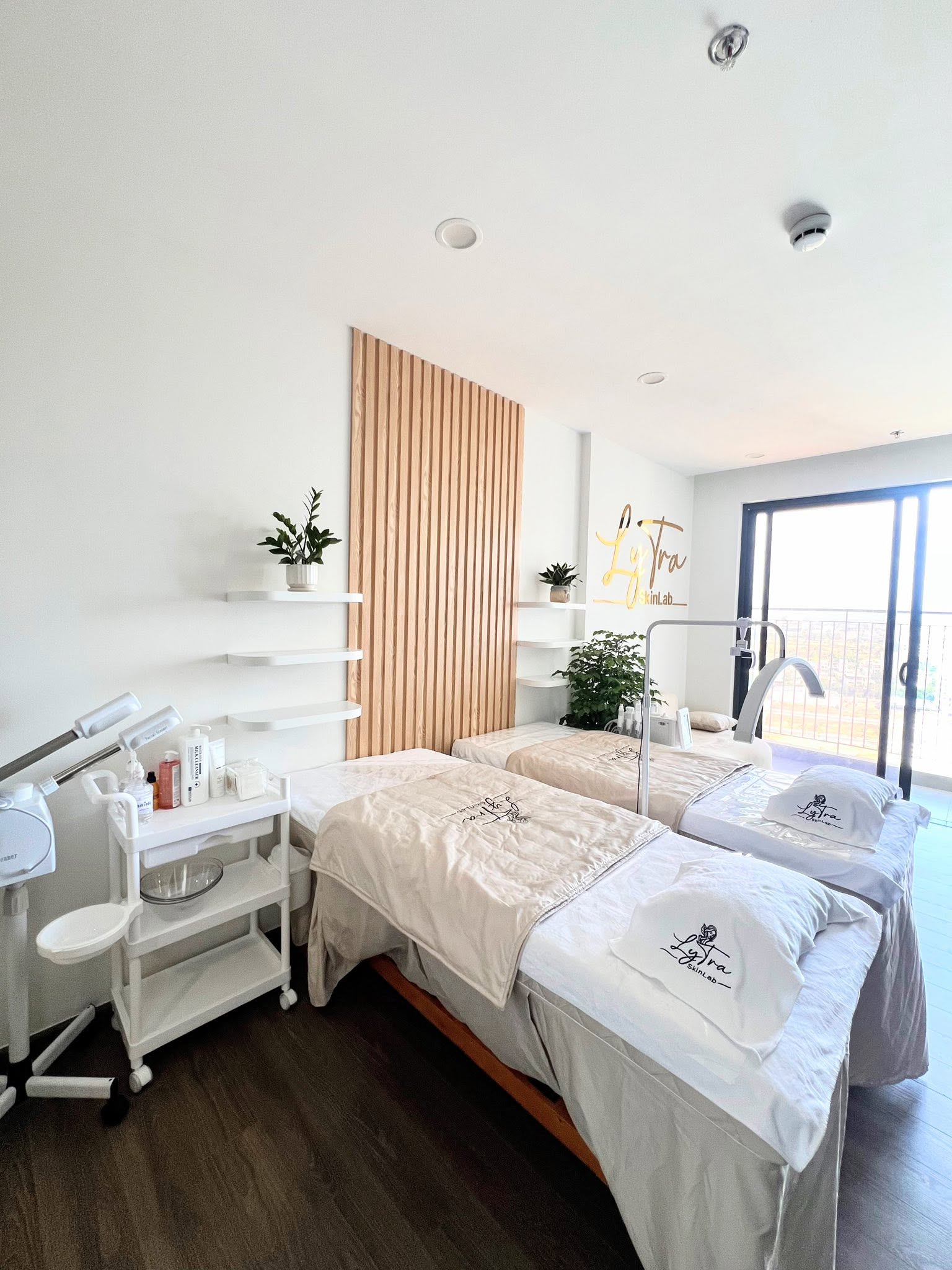 Hai giường treatment LyTra SkinLab, tường gỗ thanh và kệ sản phẩm trắng